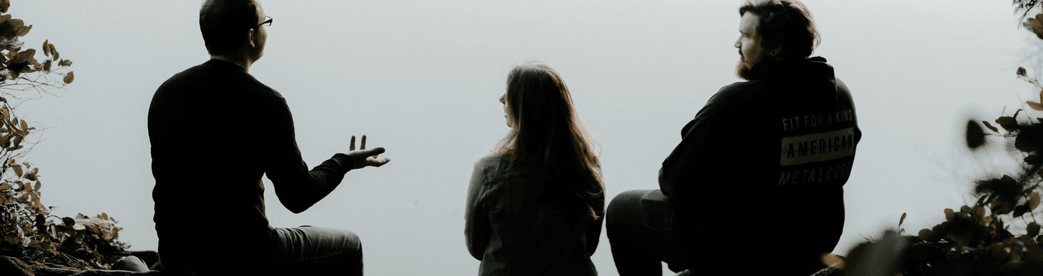 Three silhouetted people sitting by a lake in conversation, viewed from behind